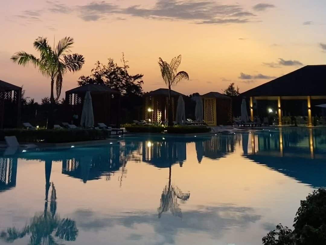 Piscine d'un hôtel béninois au crépuscule, palmiers et paillotes se reflétant dans l'eau